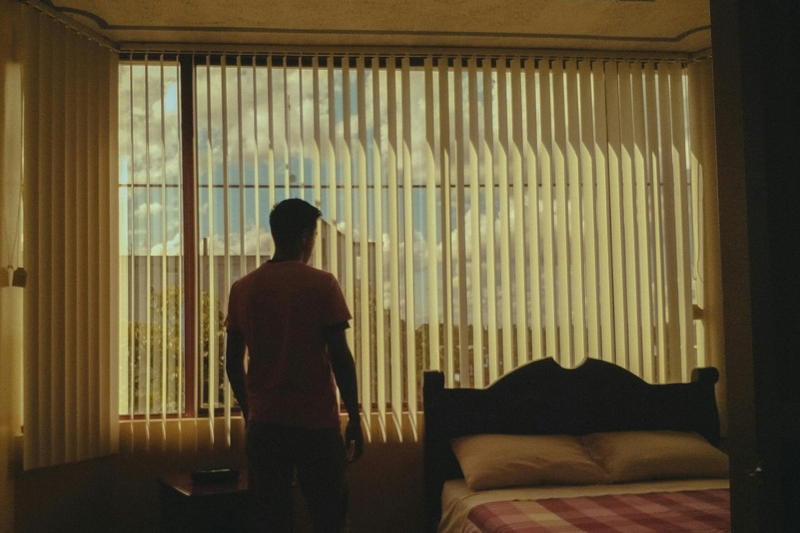 A silhouette of a man stares out a bedroom window with closed blinds, emphasizing introspection.