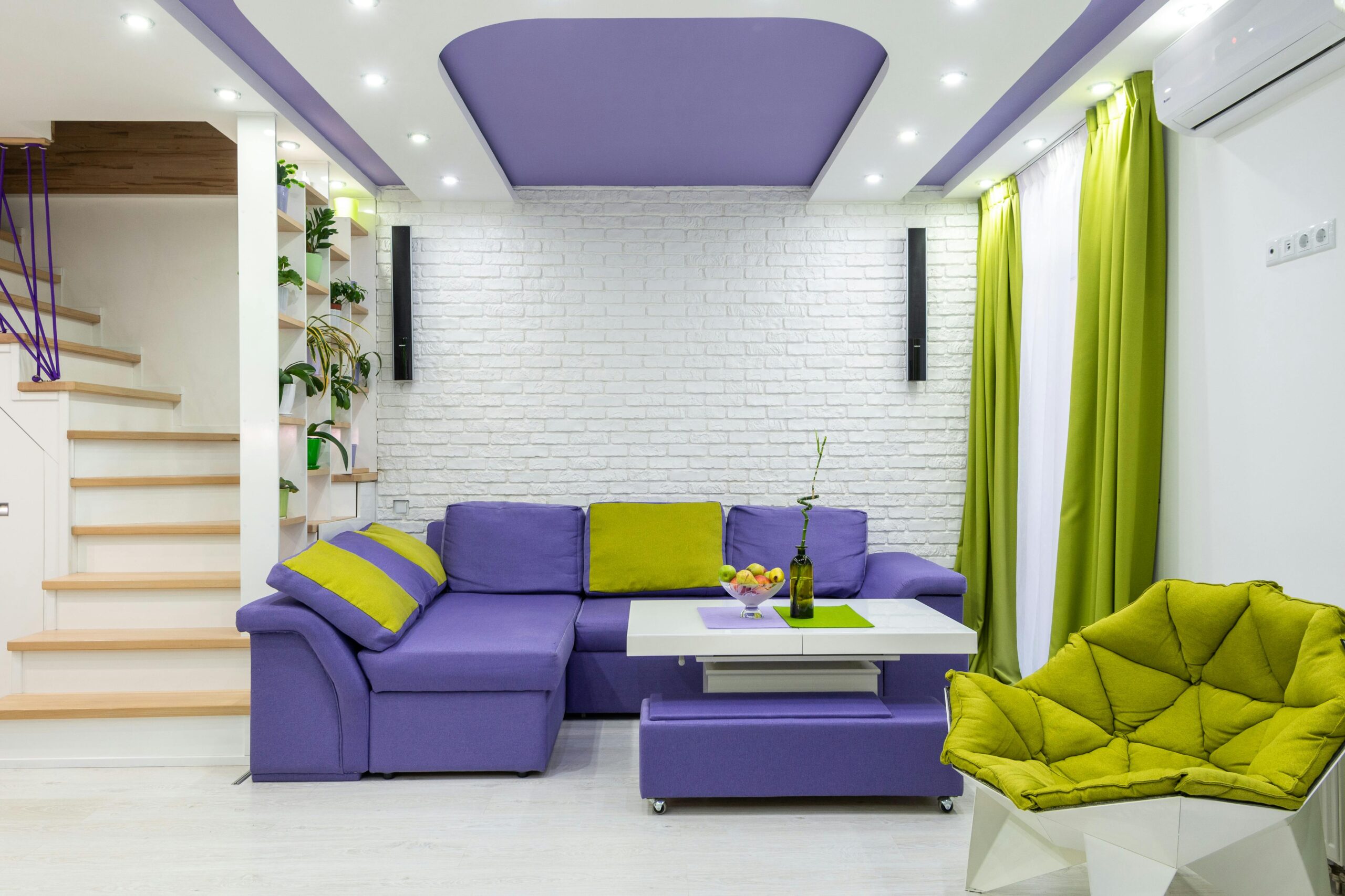 Comfortable purple and green corner sofa with coffee table and armchair placed near white brick wall in contemporary house with suspended ceiling and staircase