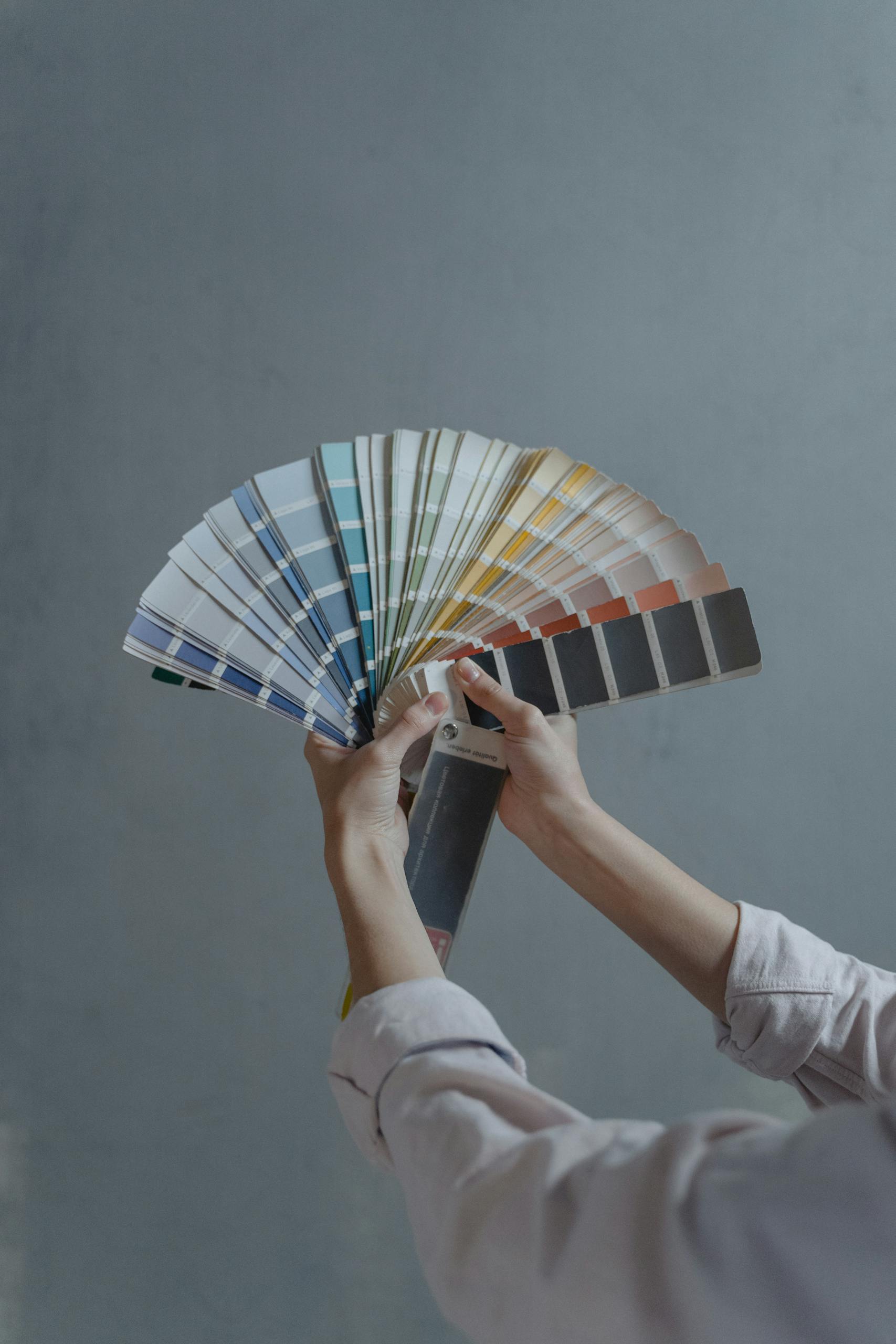 Hands displaying a fan of paint color swatches against a gray wall